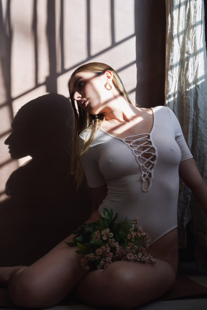 Marintia Bustos a model sits gracefully against a wall her face illuminated by sunlight filtering through window blinds casting a shadow on the wall. She wears a white lace-up bodysuit and holds a bouquet of pink flowers.